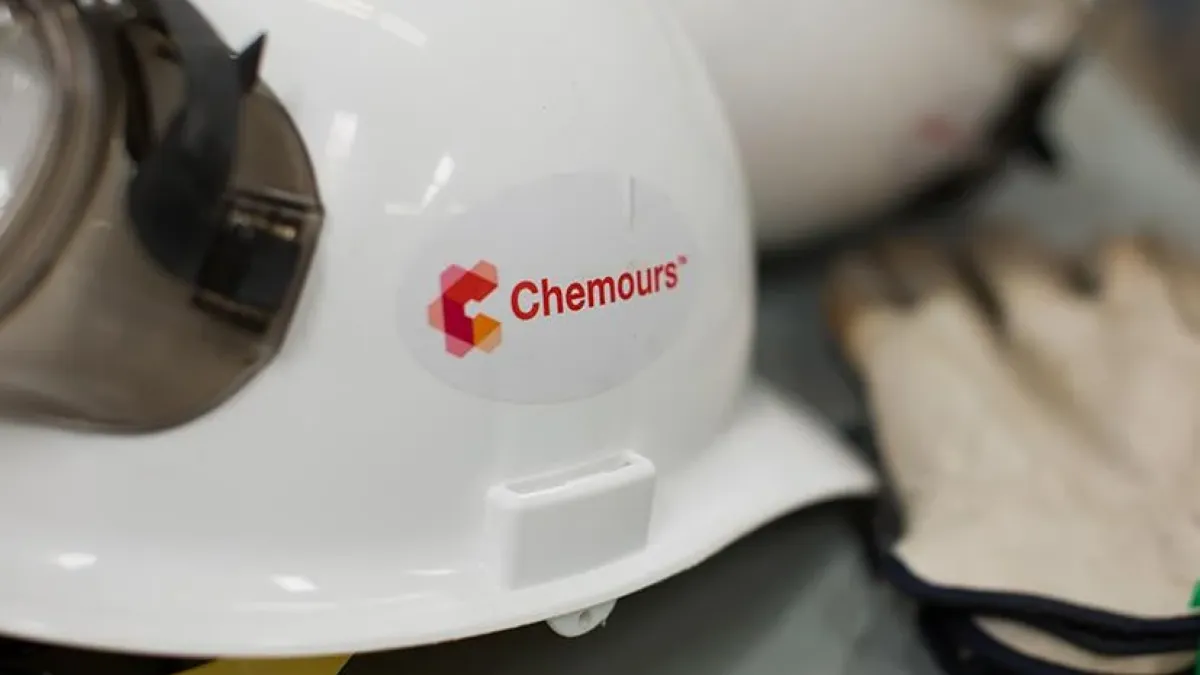 A white hard hat helmet with The Chemours Co. red and orange logo in the center, laying next to a pair of tan gloves.