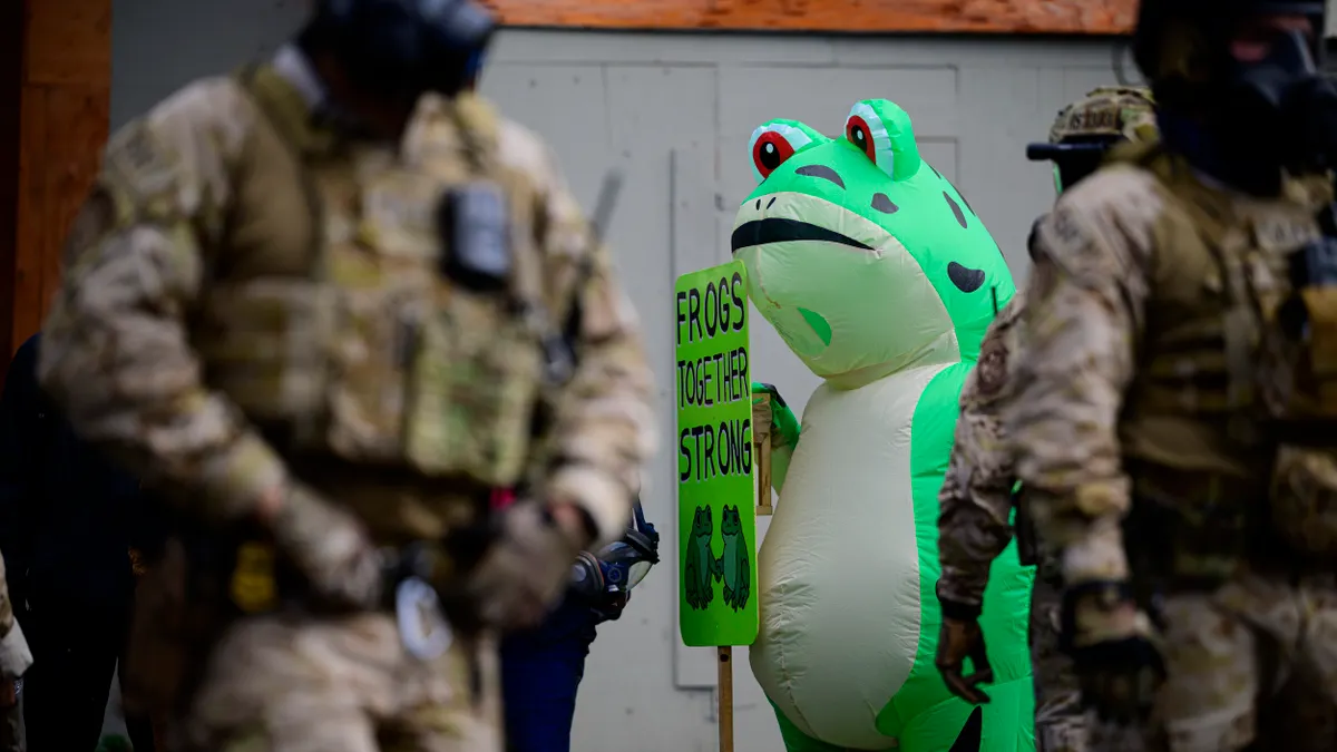 A person wearing an inflatable frog costume holding a sign that says "Frogs Together Strong," surrounded by officers wearing military gear.