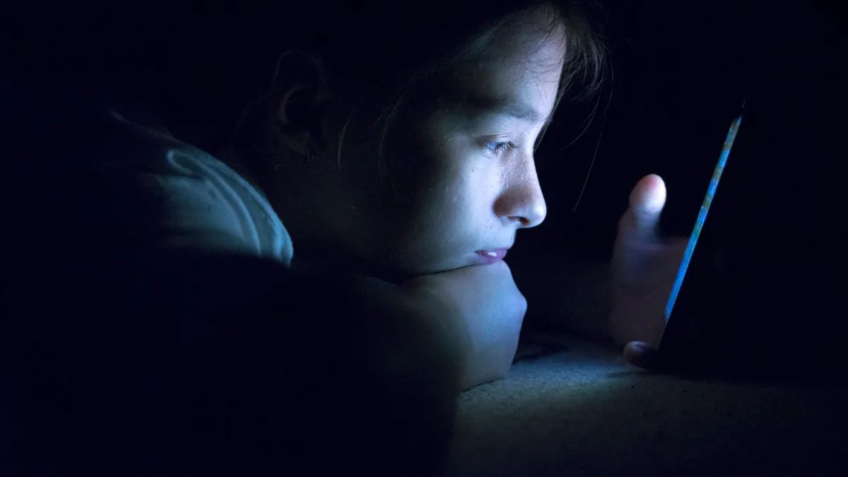 A side profile of an adolescent lying down in the dark looking at the screen of a smartphone. The screen illuminates the person's face.