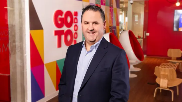 An image of a person standing and smiling. The person is wearing a navy blue suit.