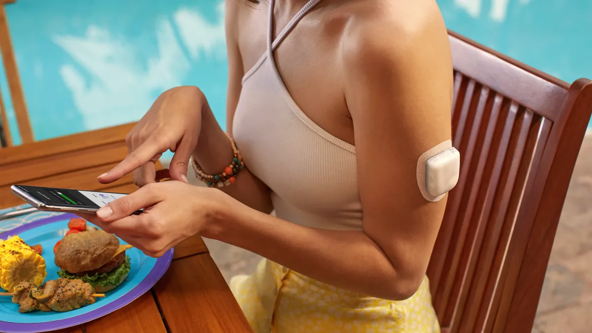 A person sits in front of a plate of food, holding a smartphone. They're wearing a patch on their left arm.