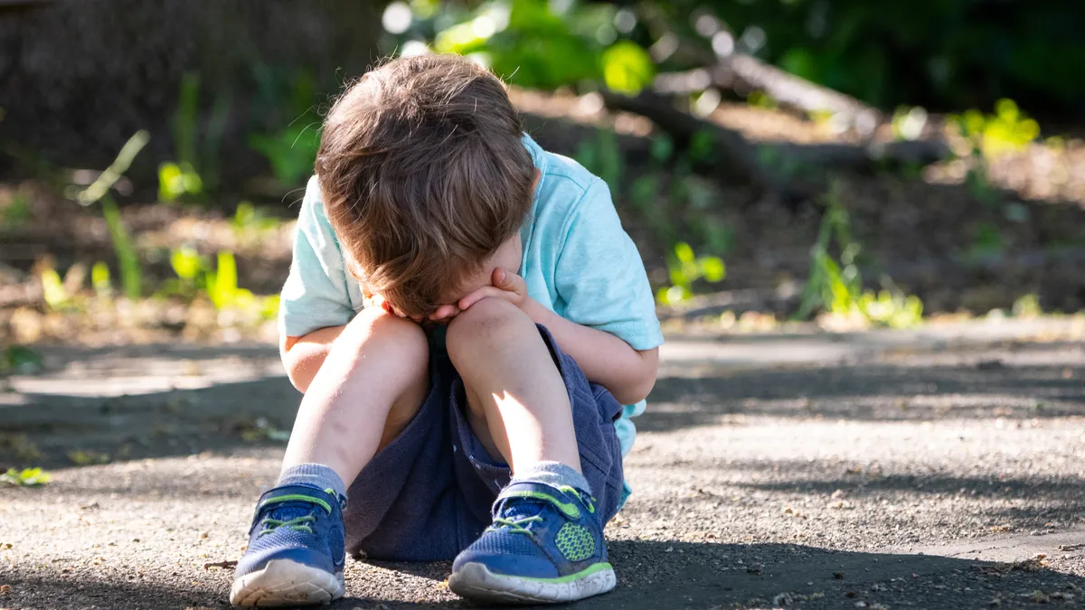 A child sits on the ground outside with their head in their hands and in their lap