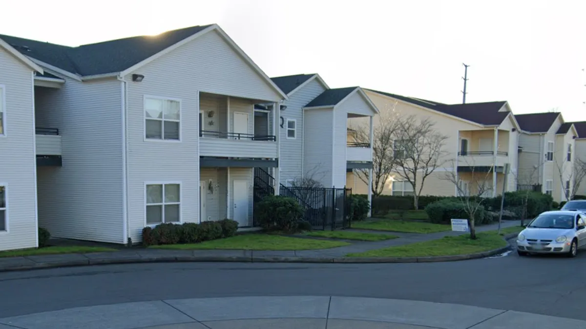 A low-rise apartment property with white siding and cars on a nearby street.