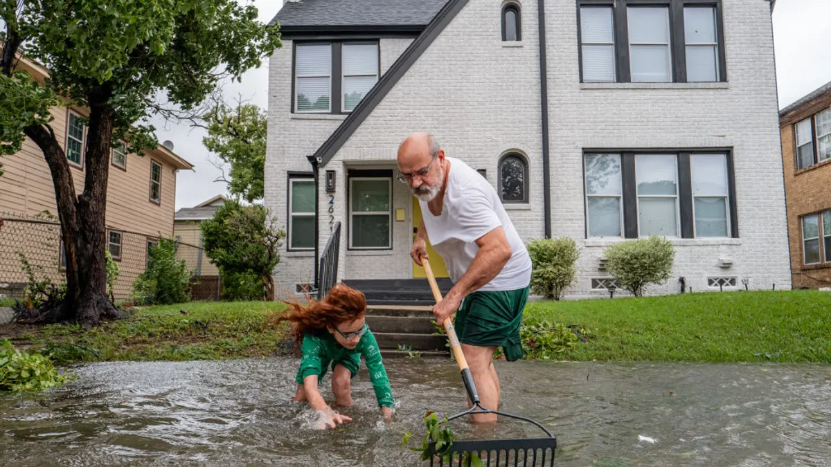 An adult holding a rake and a child stand in floodwaters outside a home.