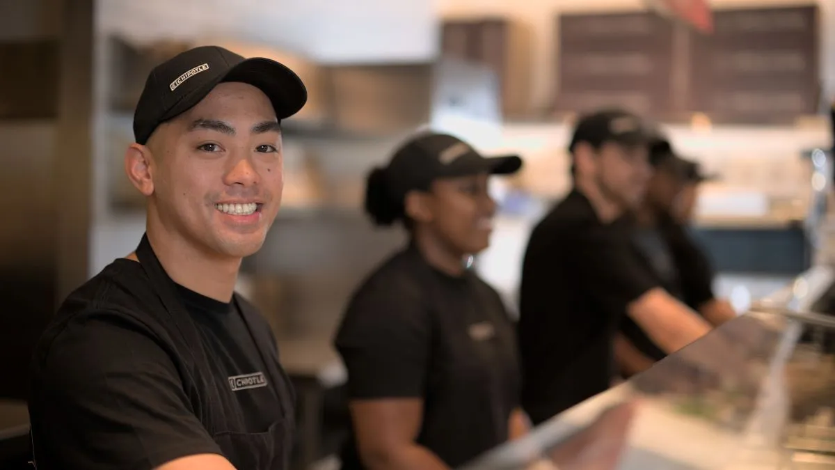 A young man wears a black hat and black apron smiles at the camera. There are three people blurred out in the background. They are standing behind a makeline.