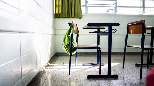 A desk and chair are pictured in a classroom that has a window. A backpack is hanging off the back of the chair.