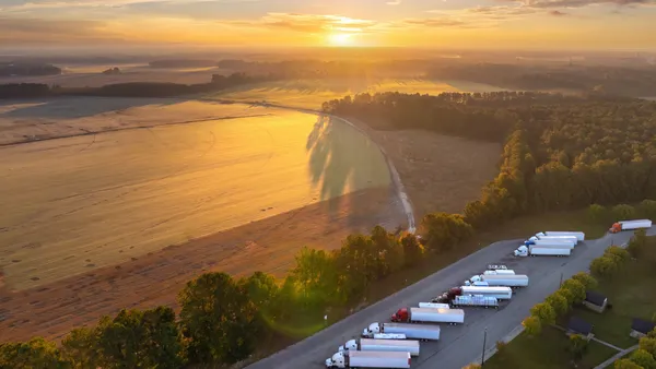 Big parking truck stop for tractor trailers rest area near busy with fields and sun in background.