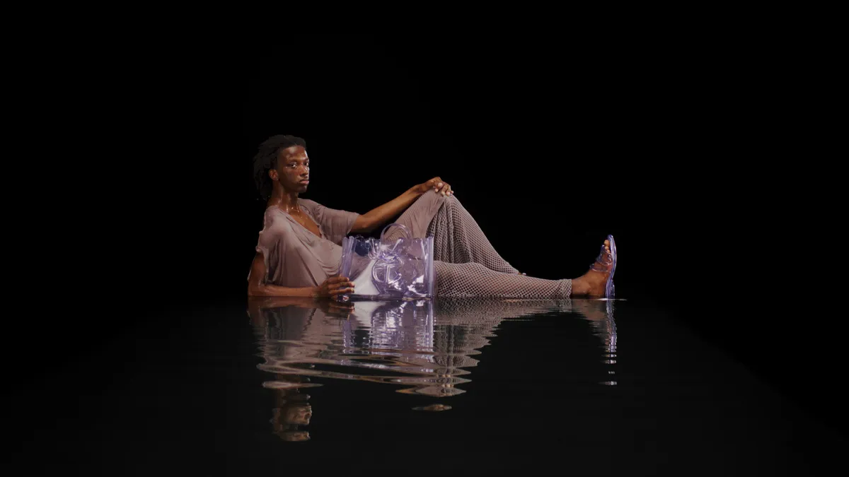 A person wears a draped brown robe and lies on a watery black background, propped up on one arm and facing the camera holing a clear plastic bag and wearing a paid of clear plastic slides.