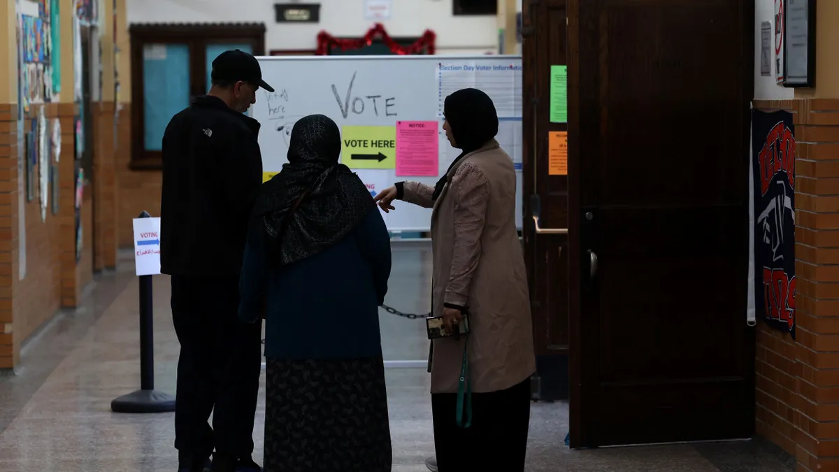 Three voters stand in front of a sign that says "vote here" with an arrow pointing to the right as they wait to cast their ballot in an elementary school.