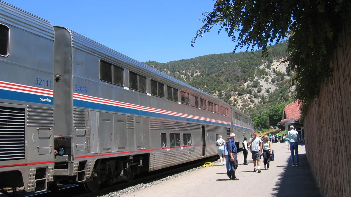 Passengers walk along a platformat a station next to a bilevel Amtrak train with hillside trees in the background.