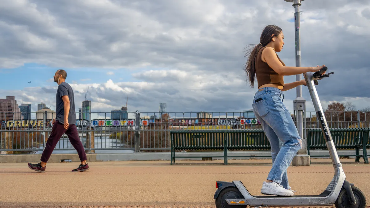 A person rides by on a Bird scooter as a pedestrian with headphones in walks in the other direction. Behind them is a waterway, benches and city buildings.