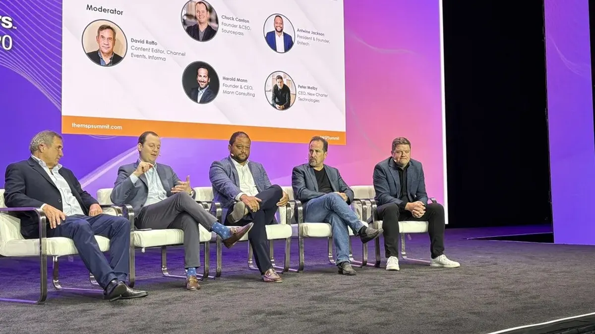 Five men sit on chairs on stage in front of a screen that shows their names and titles