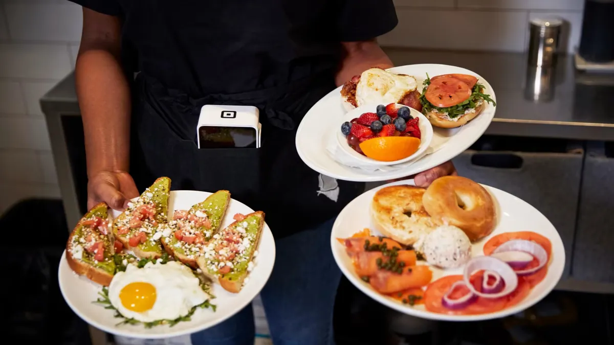 A person carrying three plates of food on their arms and hands while they have a square device wrapped around their waist.