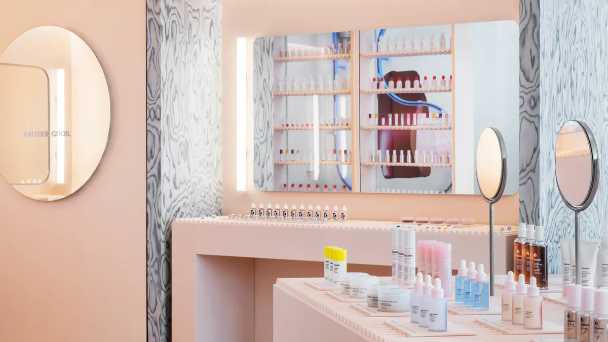 Glossier skincare and makeup products sit on pink display shelves in a pink-walled room.