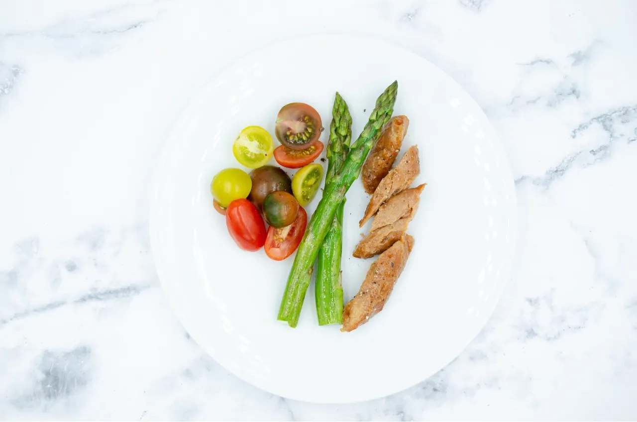 A white plate with pieces of roasted Air Chicken, two asparagus spears, and sliced colorful cherry tomatoes. The plate is on a white and grey marble surface.