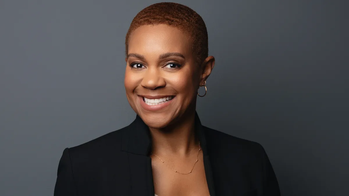 A person in a black suit jacket and short cropped hair smiles at the camera in a corporate headshot.