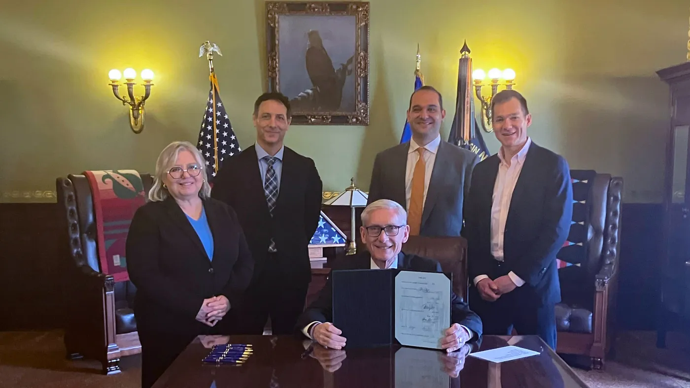 Governor Tony Evers sits at table holding up newly signed law, with others standing behind him.