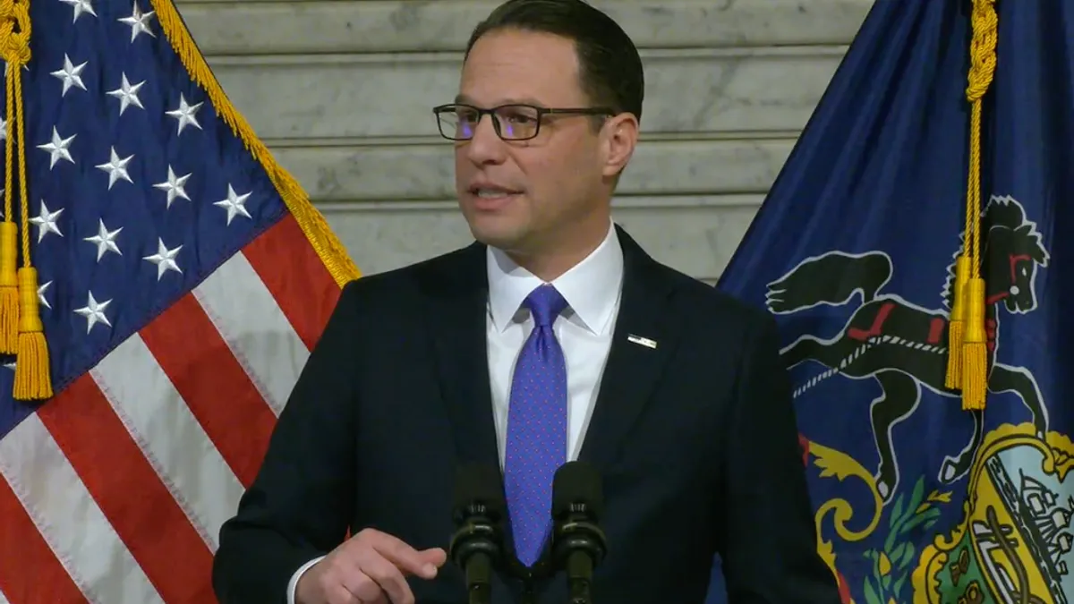 A White middle-aged man with dark hair and glasses speaks at a podium in front of the US flag and the Pennsylvania state flag.