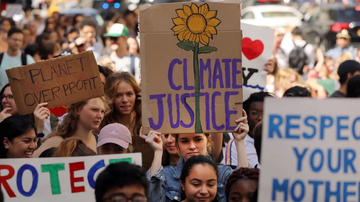 A group of young protesters marching fill the frame with signs that read "Planet Over Profit" and "Climate Justice" with a sunflower sprouting bottom to top as the "i" in climate and "t" in justice.