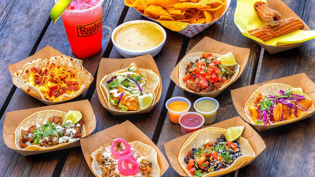 A photo of a variety of Mexican-style food on a wooden table, including a variety of tacos, nacho chips with cheese and churros. A plastic drink cup says "Rusty Taco."