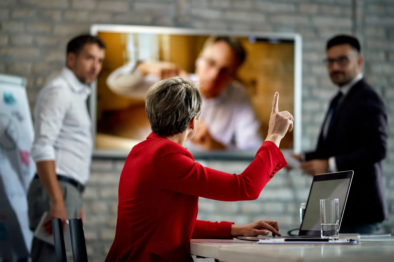 An executive wearing a bright jacket raises her hand in a meeting