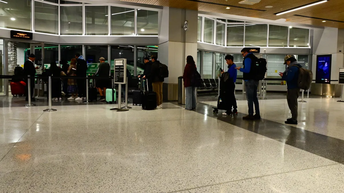 People stand in line to board a green bus seen through a glass partition.