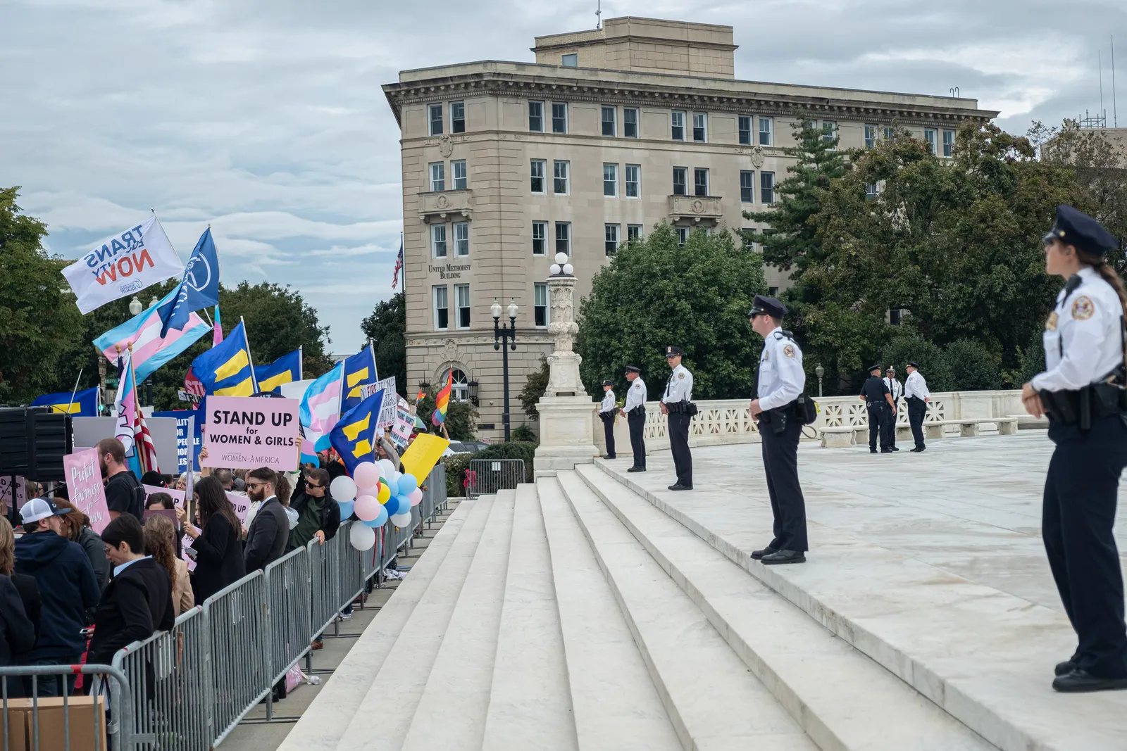 A group of protestors stand behind metal barricades while police stand parallel to the crowd.