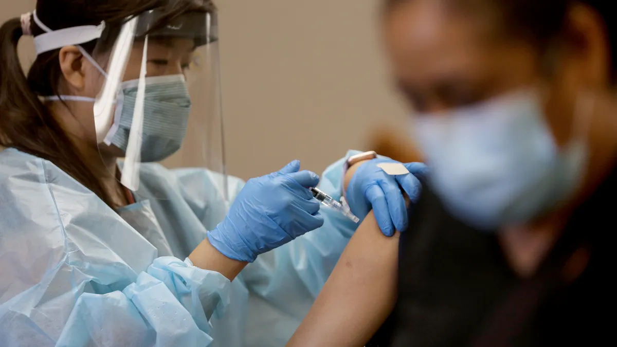 A nurse in protective equipment administers a flu shot to a woman in a mask who has been blurred out.