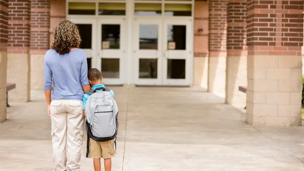 An adult stands with a student carrying a backpack outside of a school building. Their backs are to the camera.