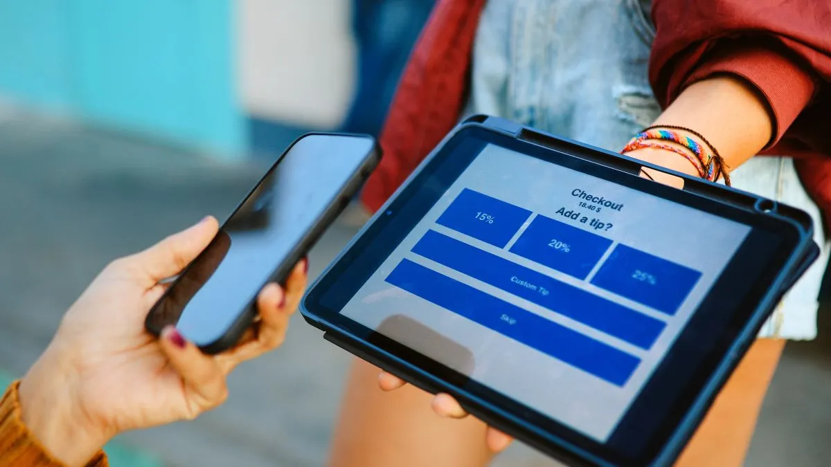 A woman holds up her smart phone to a tablet that is asking what percentage of a tip should be added.