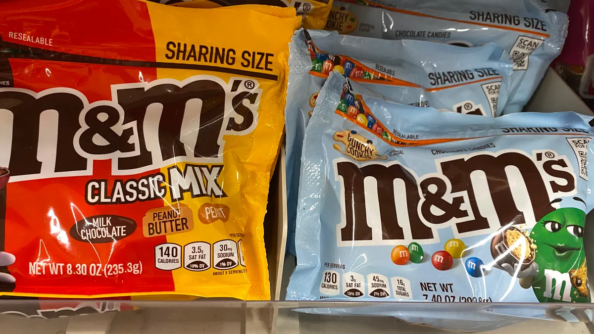 A red and yellow M&Ms Classic Mix bag and a light blue Crispy Cookie M&Ms bag on a grocery store shelf.