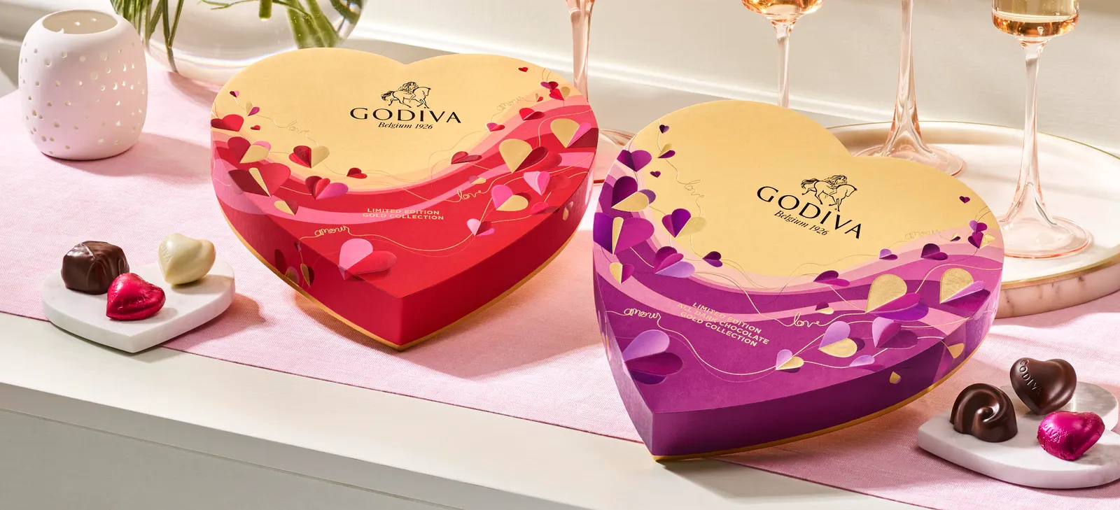 Two Godiva chocolate heart-shaped boxes --- a gold and red one on the left and a gold and purple on the right --- on a pink runner on a white table with champagne glasses. Candies are next to the boxe