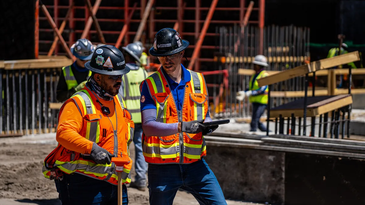 Two construction workers in orange safety vests and hard hats discuss plans at a construction site, surrounded by scaffolding and other workers in the background.