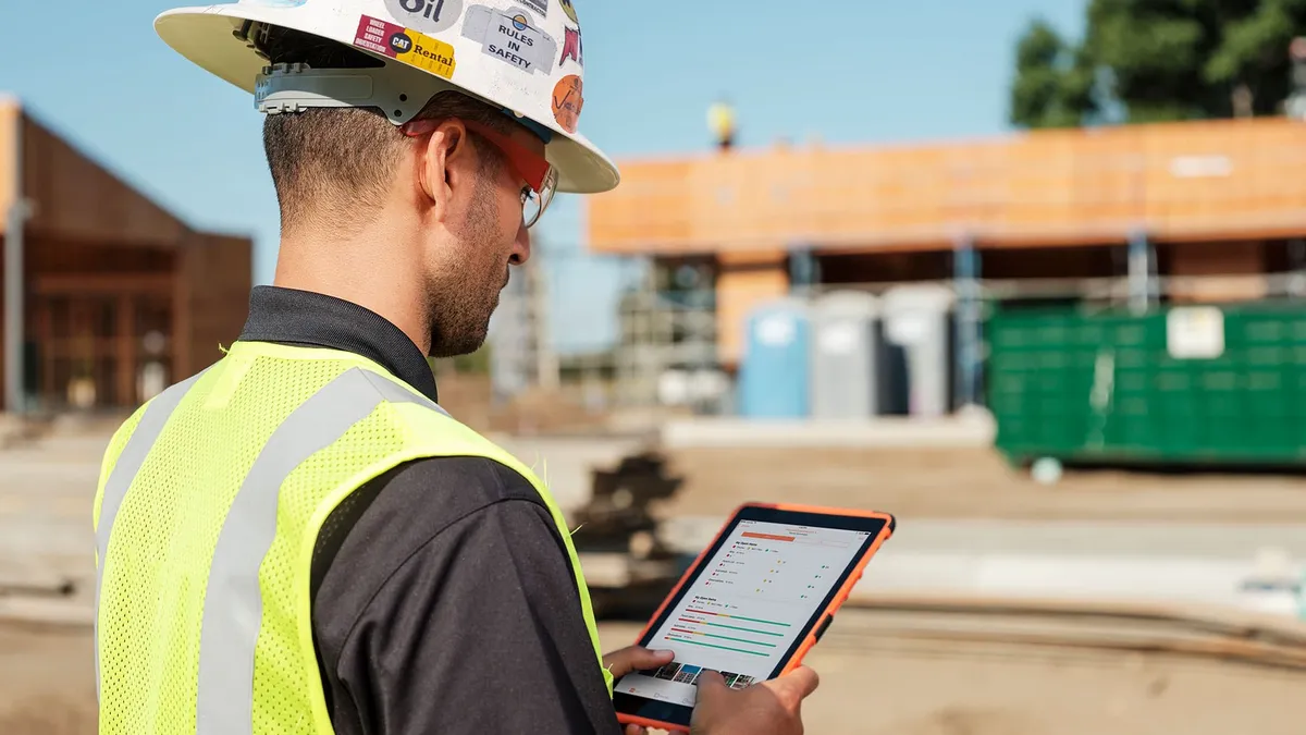 A construction worker in a white hardhat and yellow vest looks on a tablet on a jobsite.