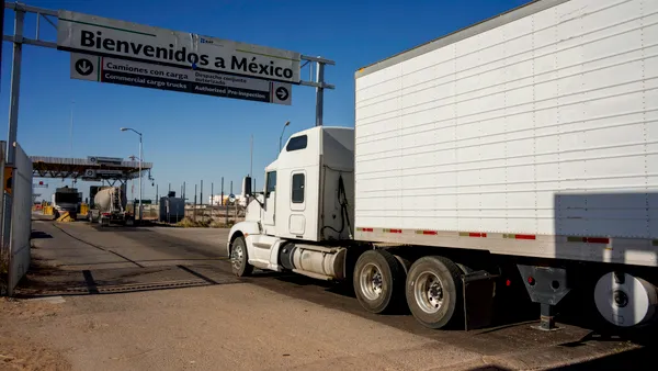 A tractor-trailer crosses an international border barrier between the U.S. and Mexico by a welcome sign in Spanish.