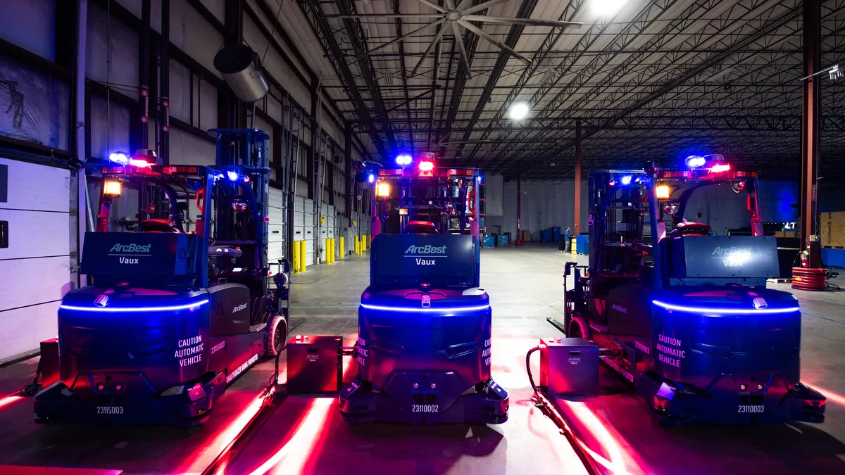 Three of ArcBest's new Vaux autonomous forklifts stand in a row with their lights illuminated in a dark warehouse.