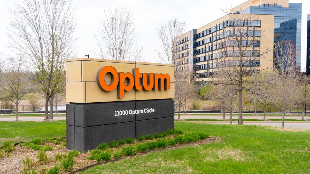 A tan and black monument sign with bold orange "Optum" lettering and the address "11000 Optum Circle" sits on a grassy lawn with low shrubs in front of a large, modern office building with glass windows and beige panels. Bare trees and a cloudy sky are in the background.