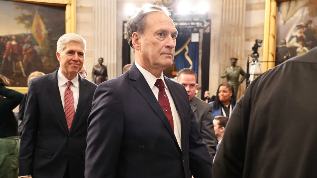 A person in a suit stands in a government building with historic artwork and statutes in the background