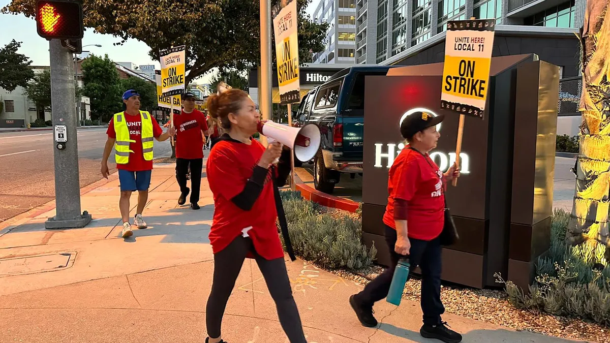 Workers in red Unite Here T-shirts hold picket signs as they march on a sidewalk in front of the Hilton Pasadena's entrance.