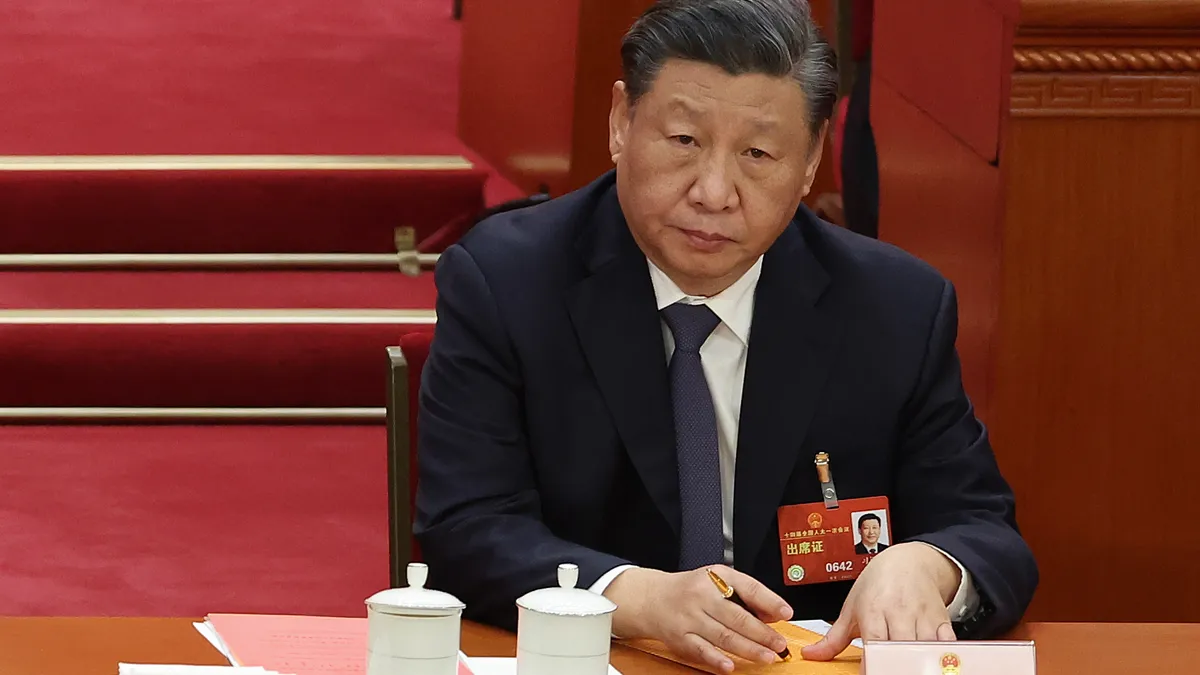 Chinese President Xi Jinping wears a suit, tie and name tag and is pictured seated at a desk and holding a pen.