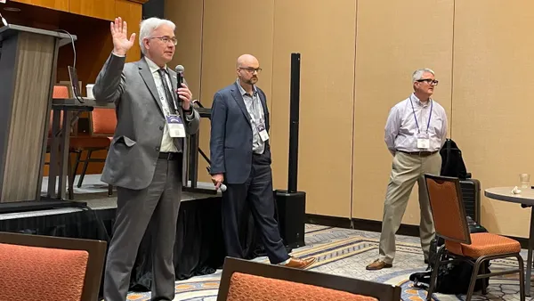 Three professional men stand at the front of a hotel ballroom, leading a discussion during an education conference.