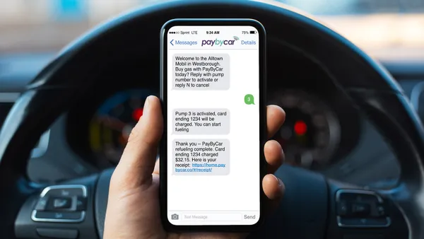 A photo of a hand holding a mobile phone while the person sits in the driver seat of a car. The screen shows text for a pay by text prompt.