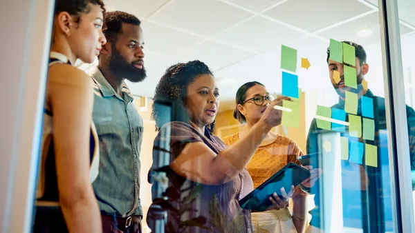 A group of diverse professionals are brainstorming ideas while standing around a glass wall with sticky notes.