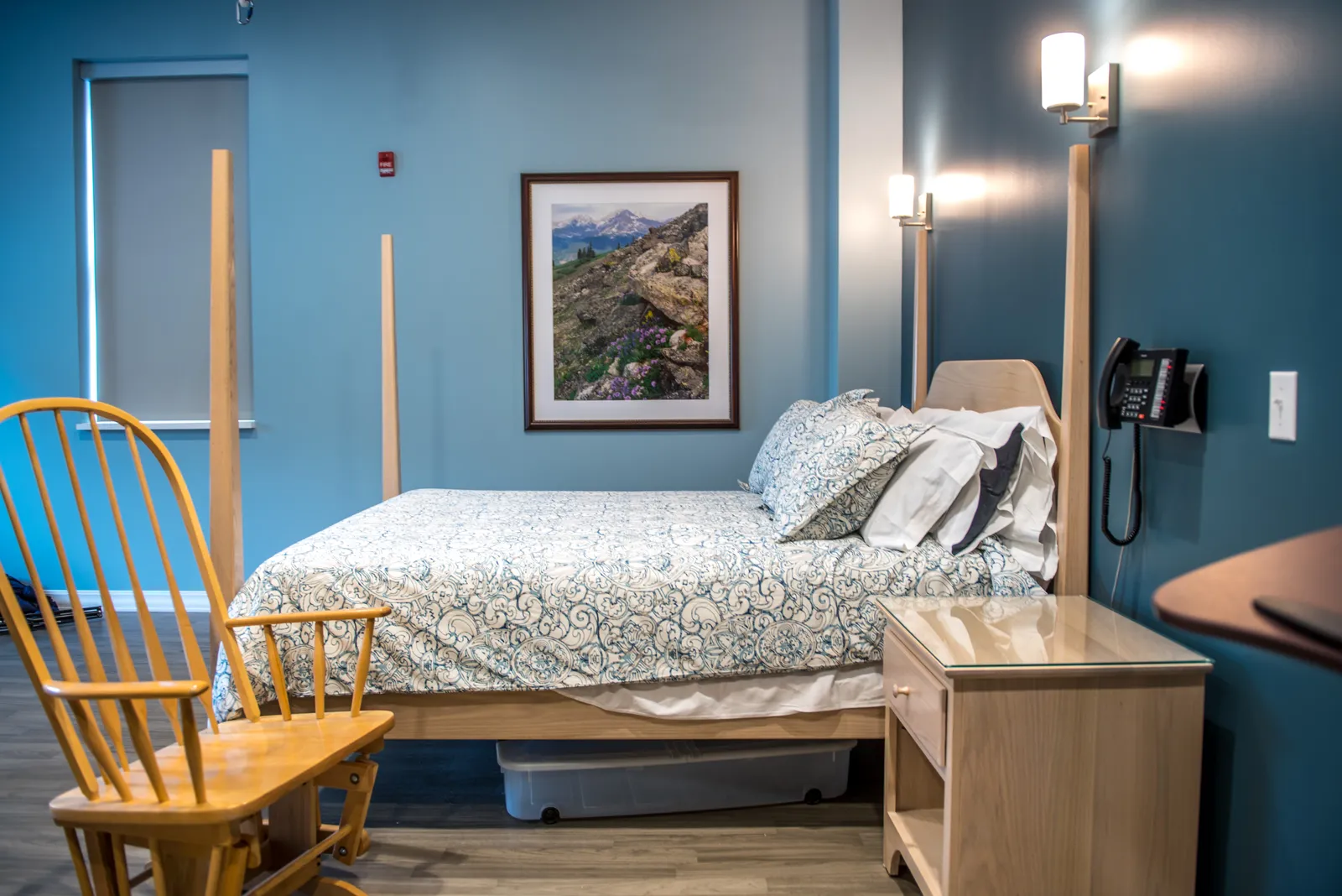 A bed sits in a calming delivery room with painted blue walls. A rocking chair is in the foreground and a photo of a mountain hangs on the wall.