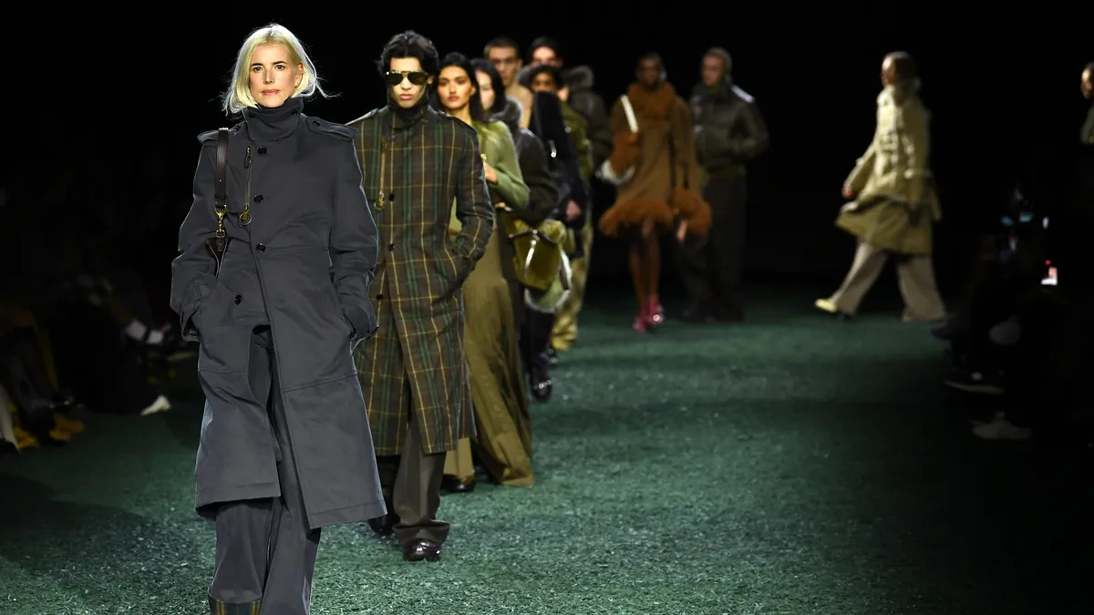 A model with blonde shoulder-length hair wears a navy coat and walks on a runway as other models follow. Spectators watch in the dark on the side.