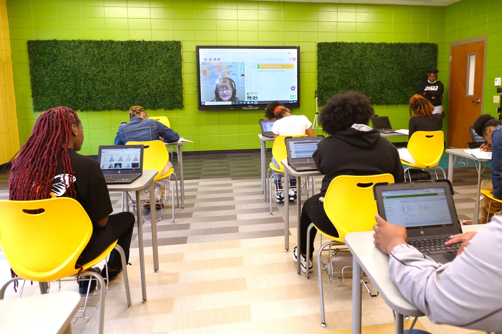 A teacher instructs remotely on a screen in front of an in-person classroom of high school students.