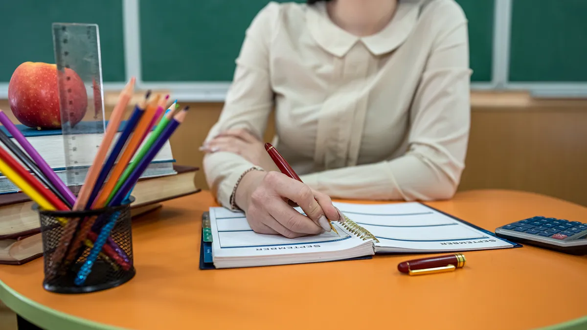 An educator sits at a desk with book, pencil, notepad and apple. Behind them is a classroom blackboard.