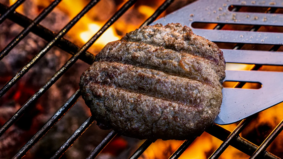 A burger made from Omeat's cultivated beef cooks on a grill. A spatula is underneath it, poised to flip it over.