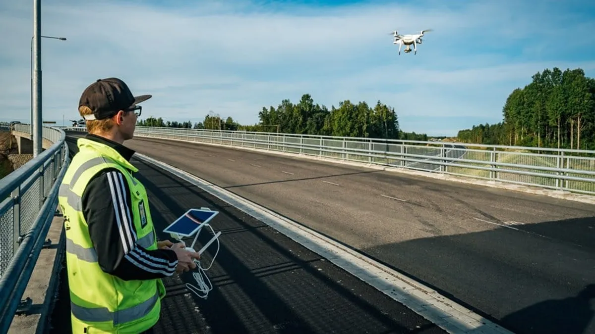 A person in a bright-green safety vest and a baseball cap pilots a small drone. It is done during the evening in fair weather.