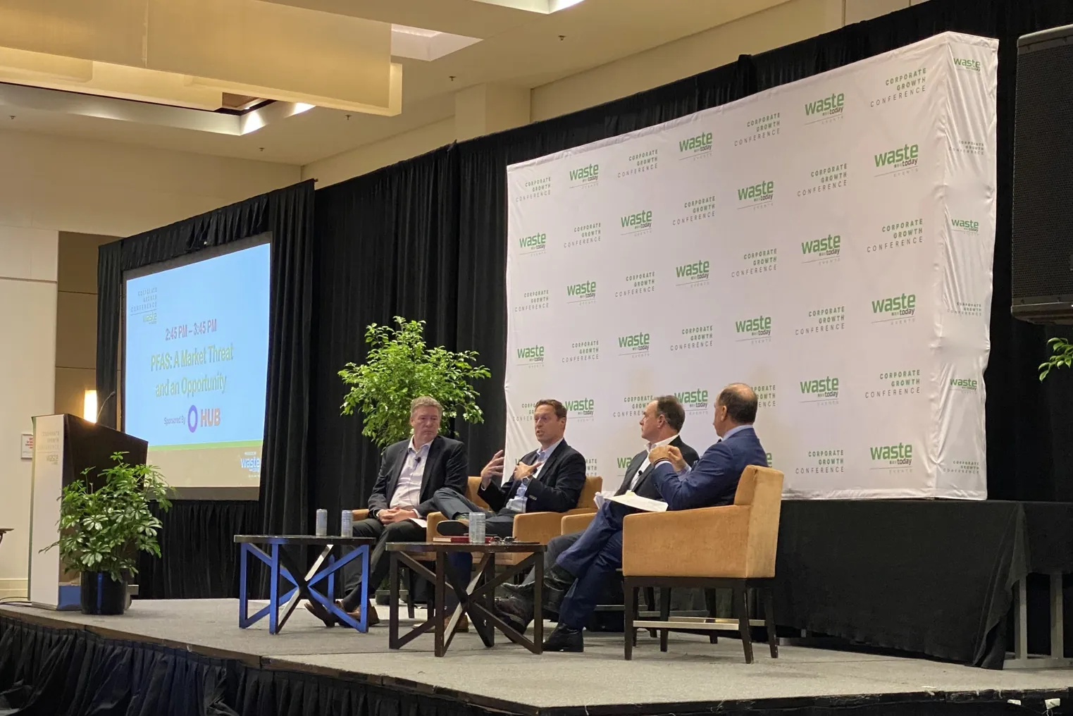 Three men are seated on a stage with a backdrop reading "Waste Today events" and "Corporate Growth Conference" behind them. One person in the center is speaking.